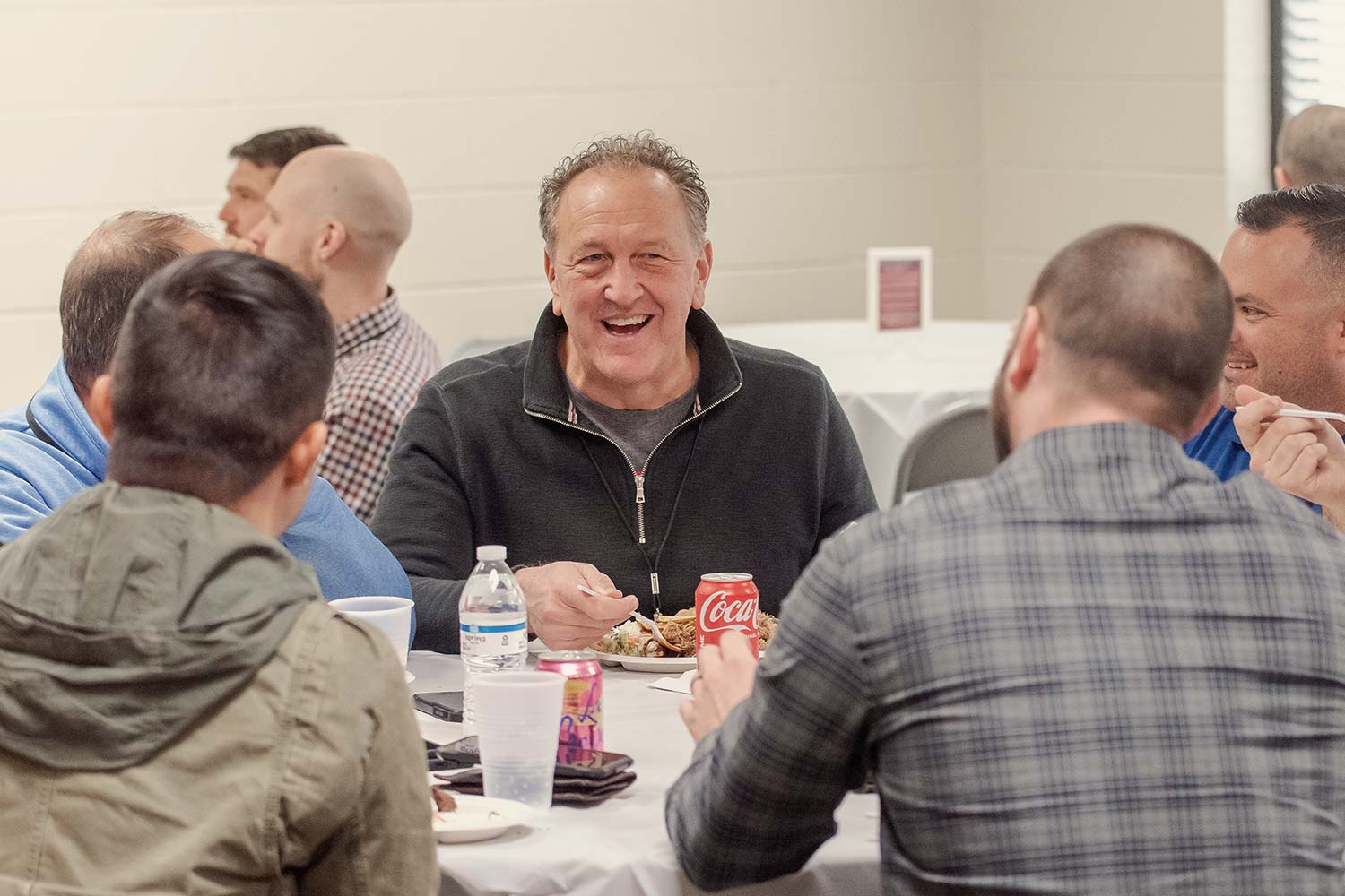 A man with a big smile sits at a table, eating and drinking a can of Coke.