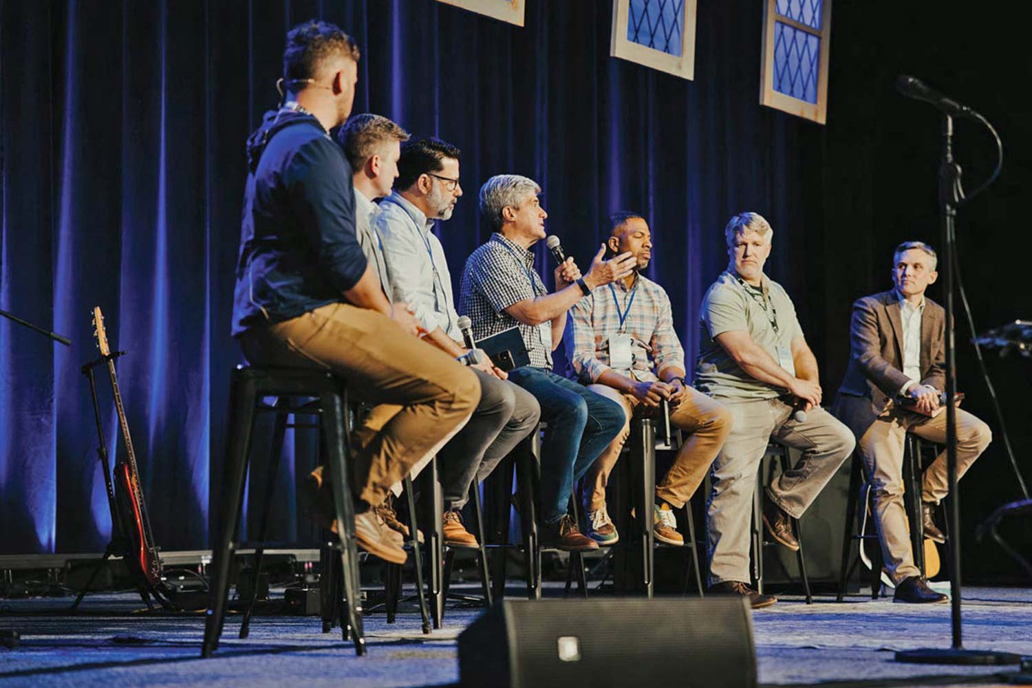 A panel of six men sit on stools on a stage, with one man speaking into a microphone.